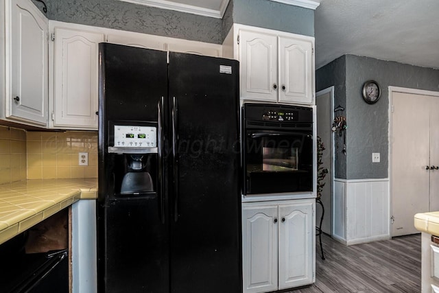 kitchen featuring tile countertops, black appliances, a wainscoted wall, and white cabinets