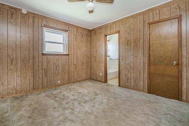 unfurnished bedroom featuring connected bathroom, wood walls, light colored carpet, and ceiling fan