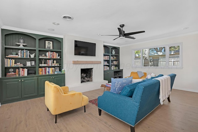 living room with crown molding, a fireplace with raised hearth, light wood-style floors, ceiling fan, and built in features