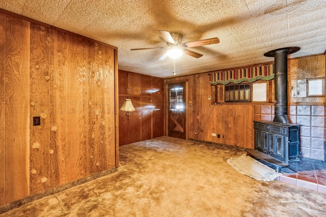 unfurnished living room with wood walls, a textured ceiling, a wood stove, carpet floors, and ceiling fan