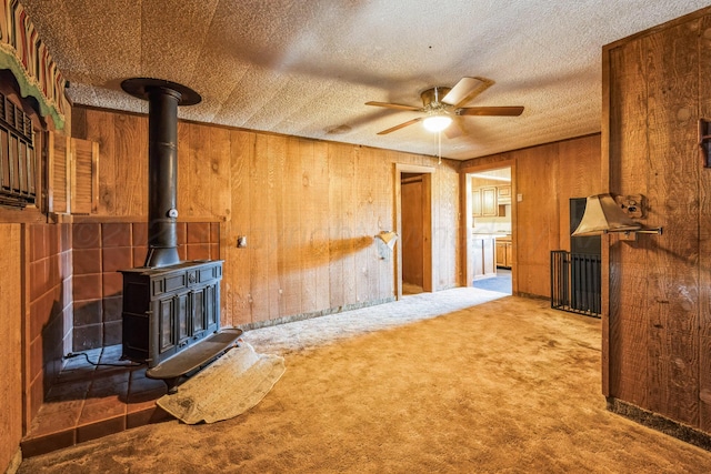 carpeted living room featuring ceiling fan, wooden walls, a textured ceiling, and a wood stove