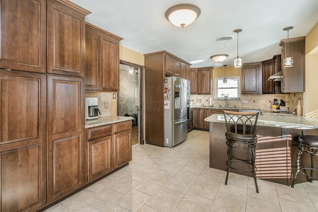 kitchen featuring stainless steel refrigerator with ice dispenser, a kitchen bar, tasteful backsplash, decorative light fixtures, and light stone countertops