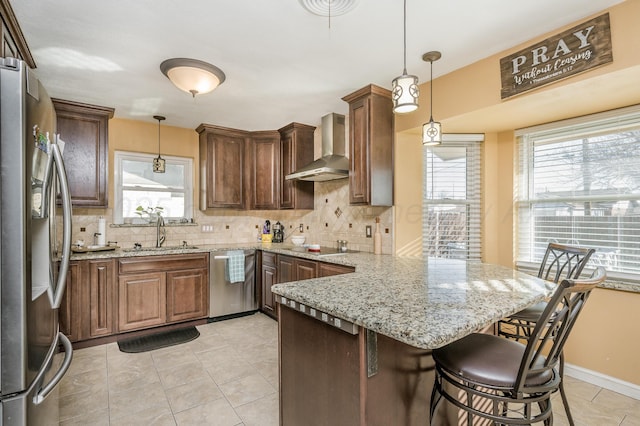 kitchen featuring wall chimney exhaust hood, sink, appliances with stainless steel finishes, a kitchen breakfast bar, and pendant lighting