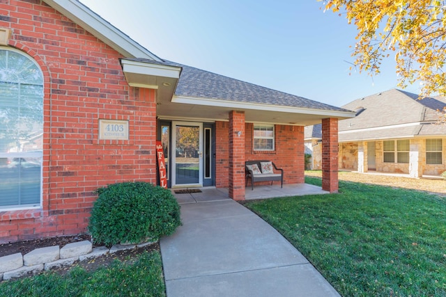 doorway to property featuring brick siding, a lawn, a shingled roof, and a porch