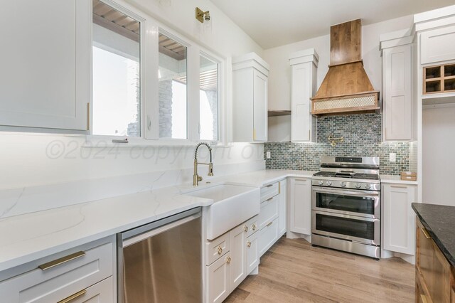 kitchen featuring light stone counters, custom exhaust hood, stainless steel appliances, white cabinetry, and a sink