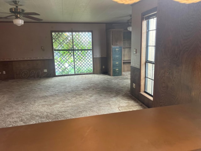 unfurnished living room with carpet flooring, plenty of natural light, ceiling fan, and wooden walls