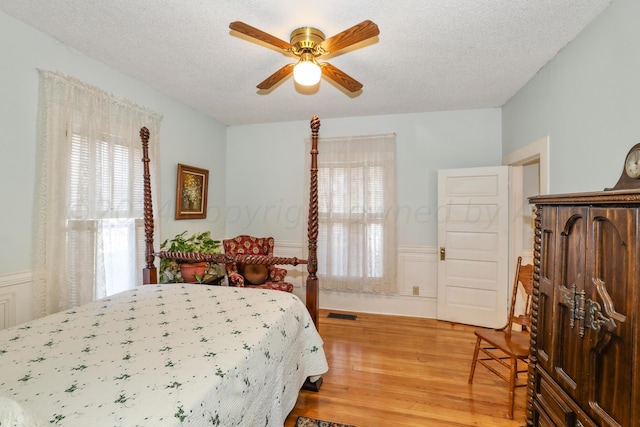 bedroom featuring multiple windows, light wood-type flooring, a wainscoted wall, a textured ceiling, and ceiling fan