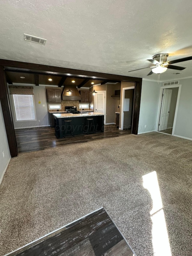 unfurnished living room with a textured ceiling, ceiling fan, visible vents, baseboards, and dark colored carpet