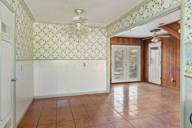 unfurnished room with ceiling fan, french doors, wallpapered walls, crown molding, and a wainscoted wall