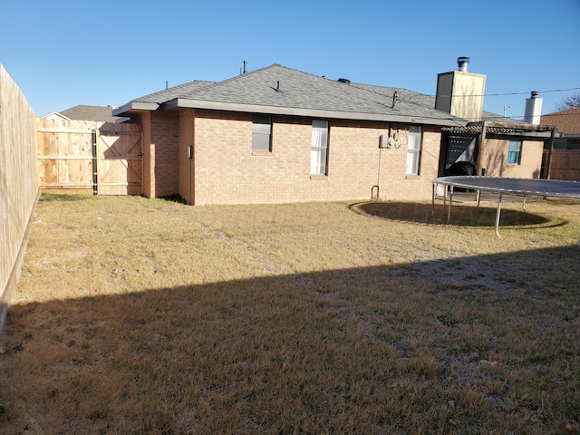 back of house featuring a lawn, a trampoline, fence, a shingled roof, and brick siding