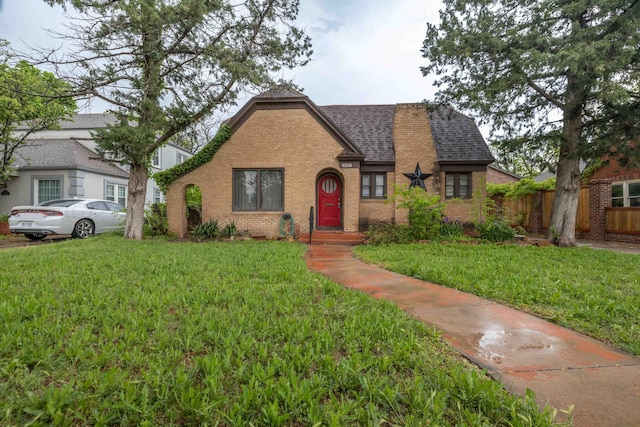 english style home featuring brick siding, fence, roof with shingles, and a front lawn