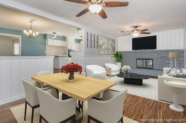 dining area with ceiling fan with notable chandelier, a brick fireplace, wainscoting, and wood finished floors