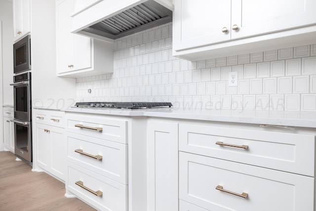 kitchen with backsplash, light wood-type flooring, custom range hood, white cabinets, and stainless steel appliances