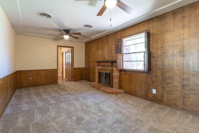 unfurnished living room featuring wooden walls, a ceiling fan, a brick fireplace, carpet flooring, and wainscoting