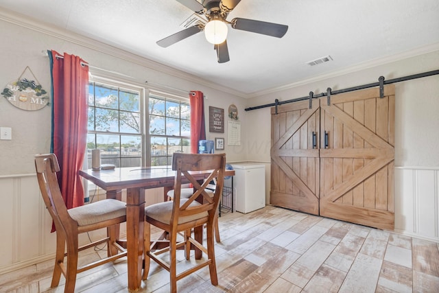 dining room with a decorative wall, a barn door, ceiling fan, ornamental molding, and light wood finished floors