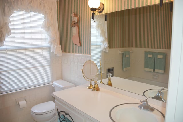 bathroom featuring tile walls, toilet, wainscoting, vanity, and wallpapered walls