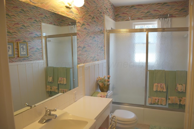 bathroom featuring a wainscoted wall, toilet, combined bath / shower with glass door, a sink, and wallpapered walls