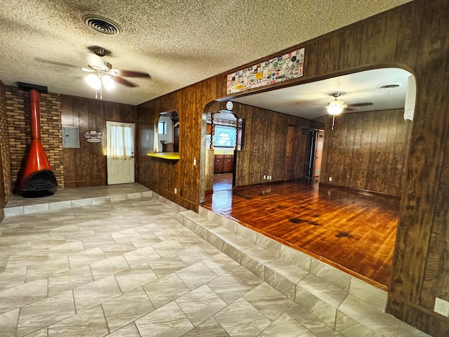 interior space with a textured ceiling, wood walls, ceiling fan, and a wood stove