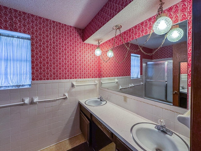 full bath featuring a sink, a shower with door, a textured ceiling, wainscoting, and wallpapered walls