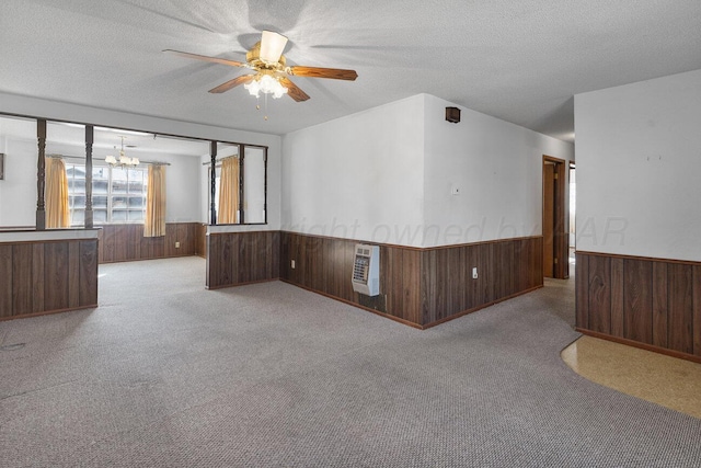 carpeted empty room featuring wooden walls, a wainscoted wall, ceiling fan with notable chandelier, heating unit, and a textured ceiling