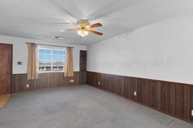 carpeted spare room featuring wooden walls, visible vents, ceiling fan, wainscoting, and a textured ceiling