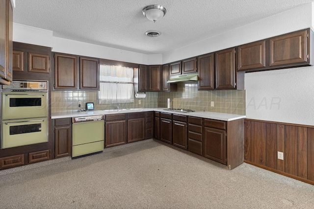 kitchen with a wainscoted wall, double wall oven, under cabinet range hood, light countertops, and dishwasher