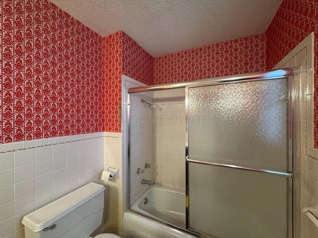 full bathroom with wallpapered walls, tile walls, a wainscoted wall, combined bath / shower with glass door, and a textured ceiling