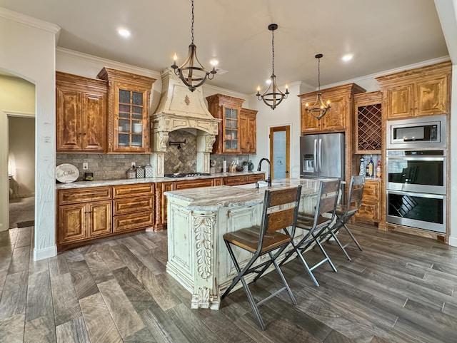 kitchen with custom exhaust hood, pendant lighting, stainless steel appliances, light stone countertops, and a kitchen island with sink