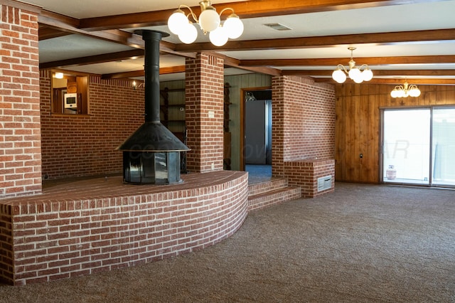 unfurnished living room featuring beamed ceiling, brick wall, a wood stove, and a notable chandelier