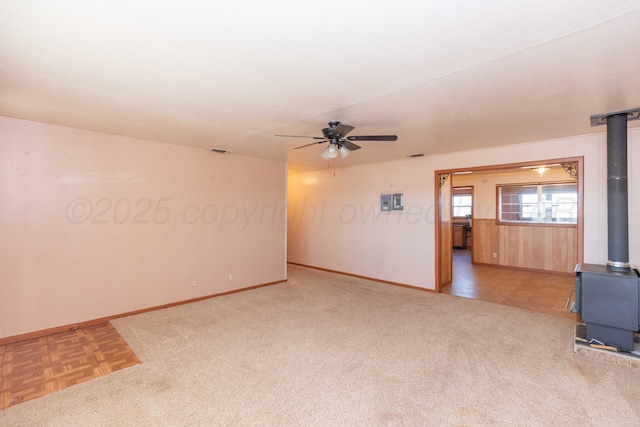 empty room with a wood stove, carpet flooring, ceiling fan, and baseboards