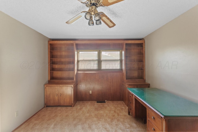 unfurnished office featuring light carpet, a textured ceiling, ceiling fan, built in desk, and wood walls