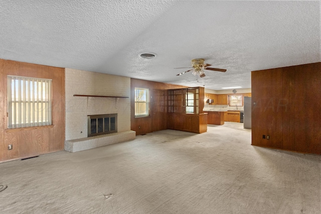 unfurnished living room featuring ceiling fan, light colored carpet, a textured ceiling, and a brick fireplace
