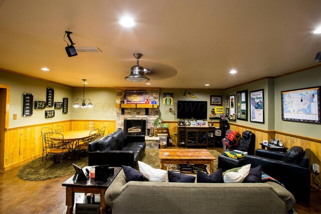 living room with a stone fireplace, wooden walls, ornamental molding, and ceiling fan with notable chandelier