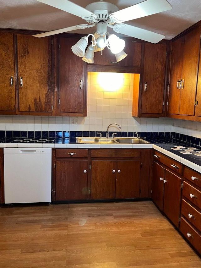kitchen featuring decorative backsplash, tile counters, light wood finished floors, a sink, and white dishwasher