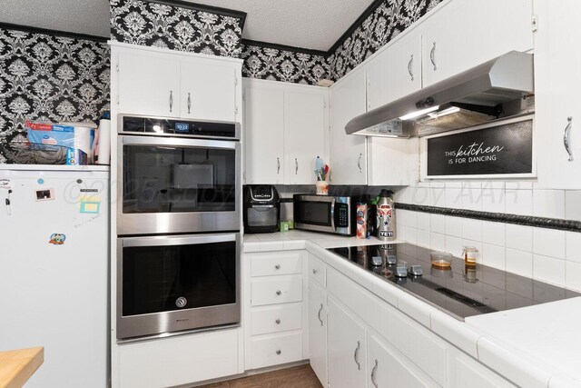 2209 N Beech Ln, Pampa, TX 79065 - kitchen with wallpapered walls, stainless steel appliances, under cabinet range hood, tile counters, and white cabinets