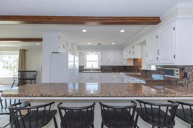 kitchen with white cabinetry, kitchen peninsula, ornamental molding, dark stone counters, and white appliances