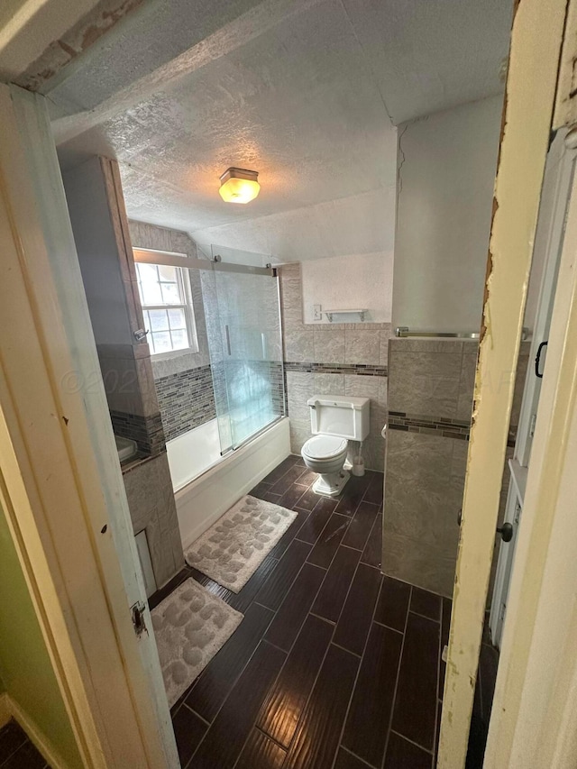 full bath featuring tile walls, wood finish floors, bath / shower combo with glass door, toilet, and a textured ceiling