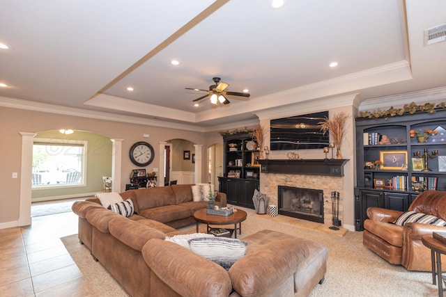 tiled living area with a ceiling fan, arched walkways, a tray ceiling, a fireplace, and decorative columns