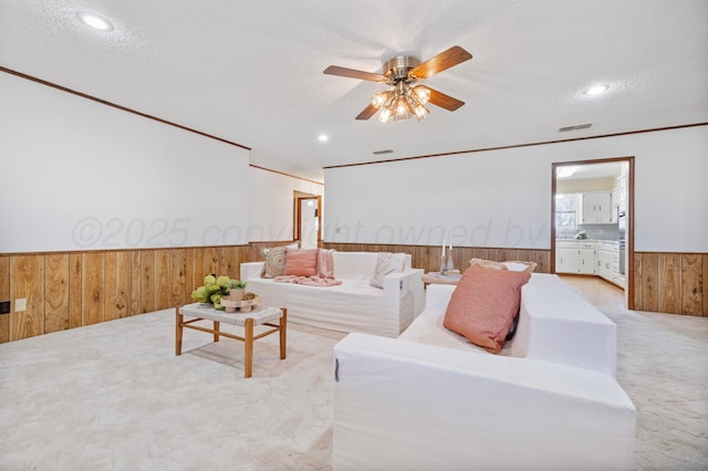 carpeted living room with wooden walls, a textured ceiling, wainscoting, visible vents, and ceiling fan