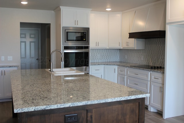 kitchen with white cabinets, premium range hood, appliances with stainless steel finishes, and an island with sink