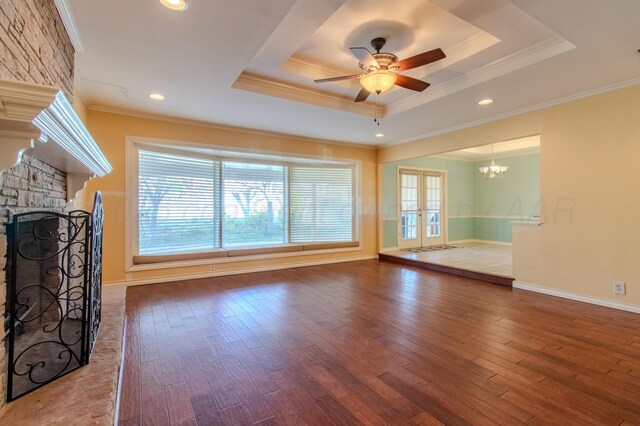 unfurnished living room featuring a tray ceiling, crown molding, wood finished floors, a stone fireplace, and ceiling fan with notable chandelier