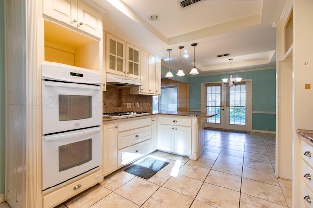 kitchen with a peninsula, white appliances, a tray ceiling, backsplash, and a notable chandelier