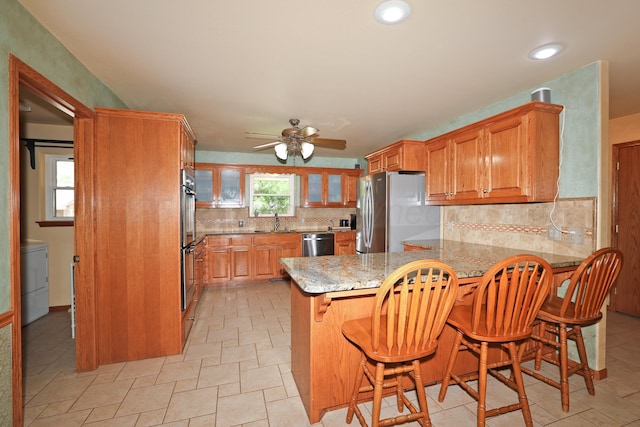1206 Olive St, Stratford, TX 79084 - kitchen featuring stainless steel appliances, a peninsula, ceiling fan, tasteful backsplash, and brown cabinets