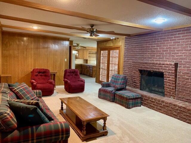 carpeted living room featuring wood walls, beamed ceiling, ceiling fan, a textured ceiling, and a brick fireplace