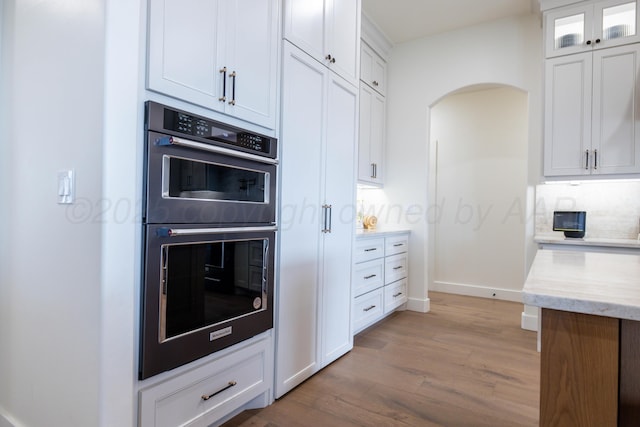 kitchen featuring arched walkways, light wood-type flooring, glass insert cabinets, multiple ovens, and white cabinetry