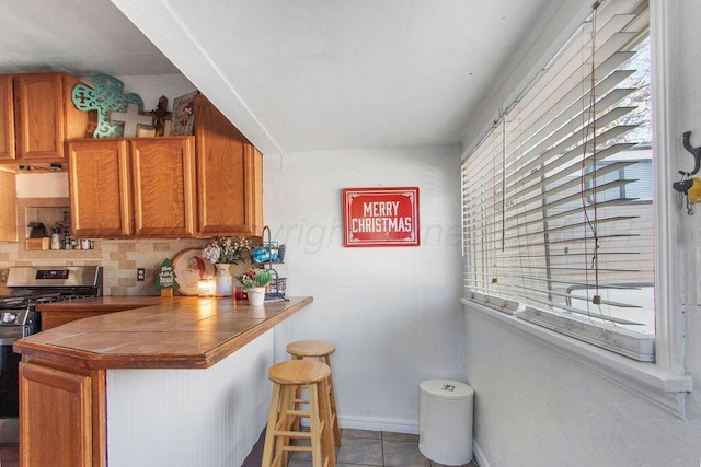 kitchen featuring tile countertops, stainless steel gas range oven, brown cabinets, and decorative backsplash