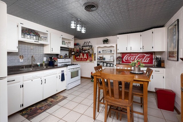 kitchen featuring white gas stove, light tile patterned flooring, sink, hanging light fixtures, and white cabinets