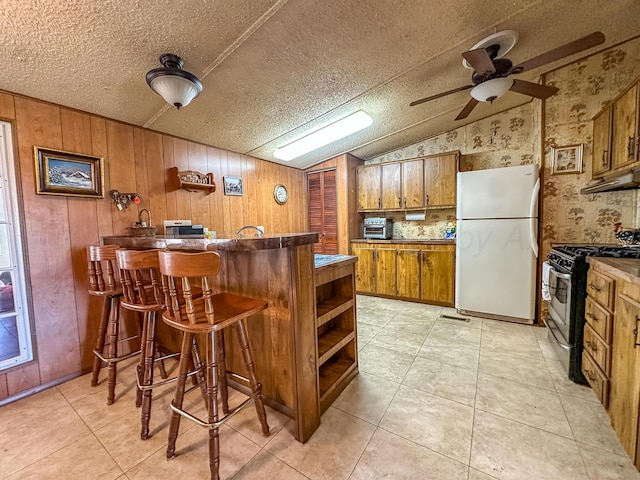 311 Jennie Ln, Borger, TX 79007 - kitchen with freestanding refrigerator, stainless steel range with gas cooktop, brown cabinetry, vaulted ceiling, and a ceiling fan