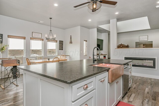 kitchen with wood finished floors, ceiling fan with notable chandelier, an island with sink, a glass covered fireplace, and visible vents