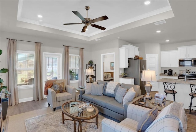 living room with sink, a tray ceiling, ceiling fan, ornamental molding, and light hardwood / wood-style flooring